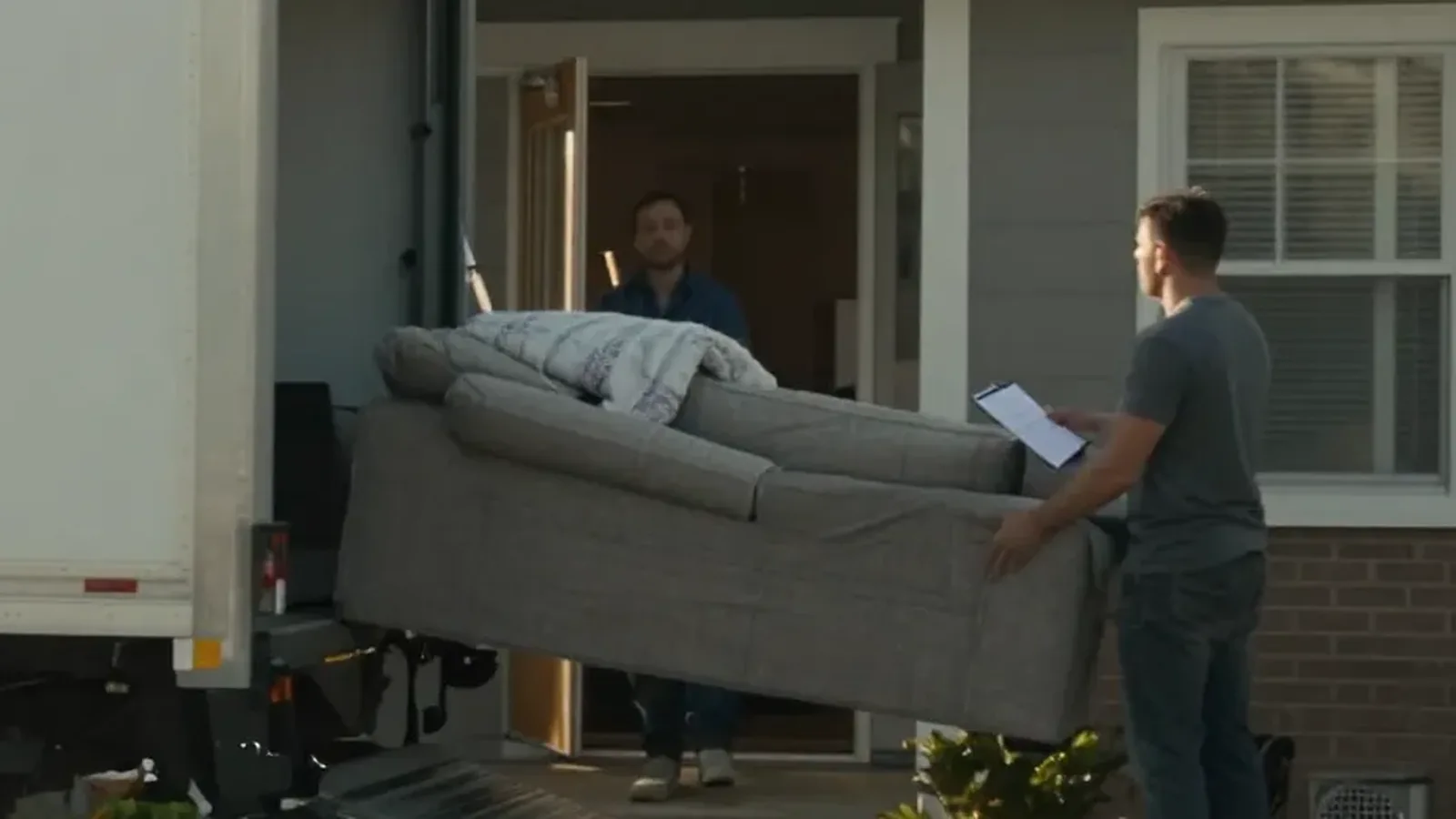 Two movers carrying a grey sofa from a box truck to a front door, one holding a clipboard
