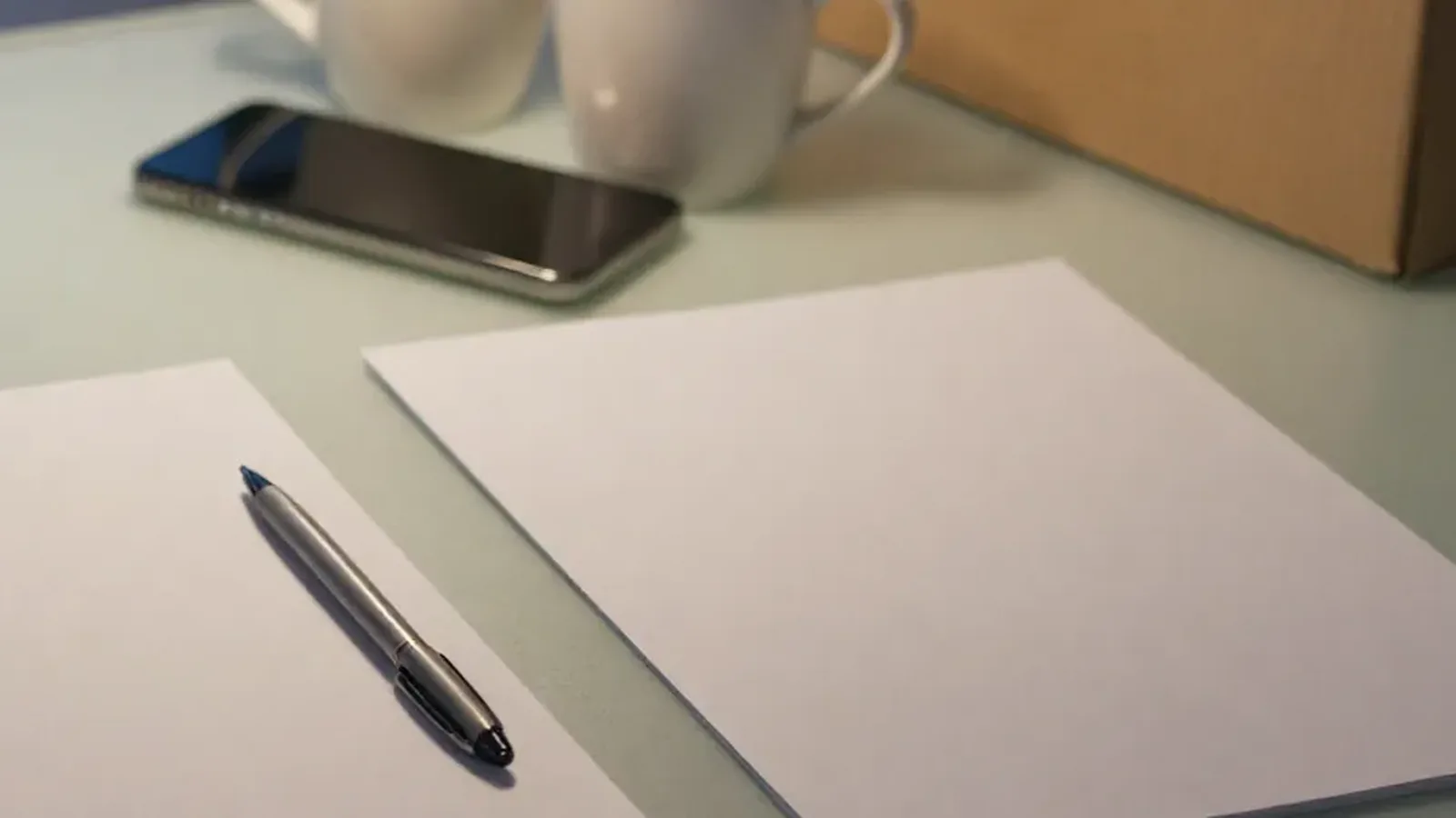 Two blank sheets of paper and a pen on a table, beside a phone and two mugs